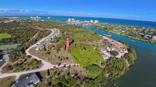 Aerial view of the red Jupiter Inlet Lighthouse surrounded by lush greenery and waterways, contrasting with the blue ocean, Jupiter, Florida, United States.