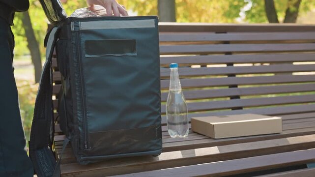 contactless food delivery on bench, insulated backpack beside bottled water and cardboard box, courier reaching for order in leafy park, wooden slats, warm autumn light, casual uniform and calm urban