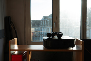 Game controller resting on top of a projector on a wooden shelf in sunlight. Casual home interior scene showing gaming setup. Concept of home, technology, gaming, leisure, lifestyle, entertainment.