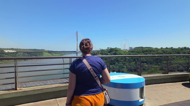 Woman tourist observes the Integration Bridge and distant Brazil-Paraguay landscape from Argentina's tri-border viewpoint at Hito Tres Fronteras
