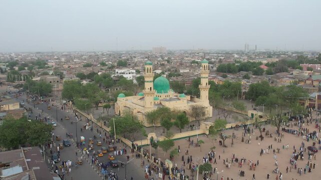 Aerial view of the Emir Palace Road Mosque, with its distinctive green dome and minarets, surrounded by bustling streets and a gathering of people, Kano, Nigeria.