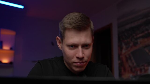 White Male Vlogger At Laptop Monitor, Dim Purple Studio Lighting And Desk Lamp, CloseUp Facial Focus, Serious Expression, Speaking Directly To Camera, Home Recording Atmosphere, Softbox Highlights,