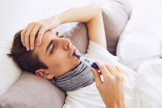 Close-up of sick man with thermometer in mouth resting on bed at home