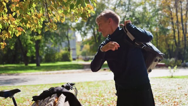 caucasian courier packing insulated bag near bicycle in autumn park, loading food parcels into thermal backpack beside bike, sunlight filtering through leaves, focused expression preparing