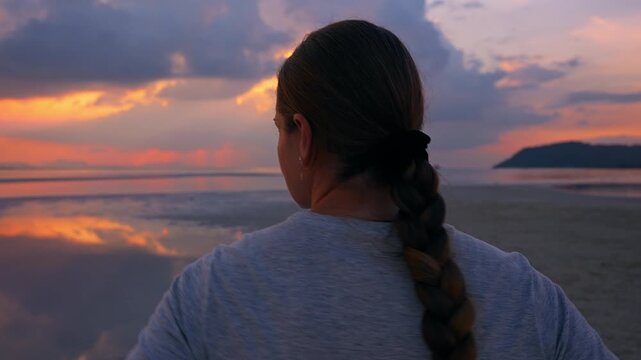 Rear Follow Shot of Woman Walking across Wet Sandflat toward Calm Sea during Low Tide with Soft Pastel Sunset Sky and Subtle Reflections, Quiet Awareness Concept