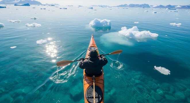 A kayaker paddles through crystal-clear Arctic waters filled with floating ice fragments toward a sunlit horizon.