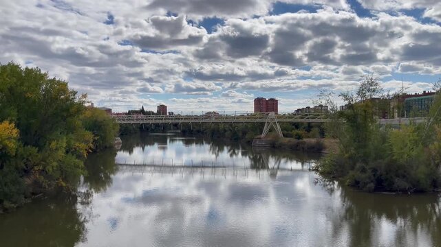 Pisuerga River flowing through Valladolid, Spain, with a modern pedestrian bridge spanning the water. Trees line the riverbanks, and residential buildings in the background under a dramatic cloudy sky