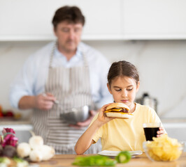 Little daughter eats meat burger and drinks carbonated drink in the kitchen. In background, father prepares healthy vegetable salad
