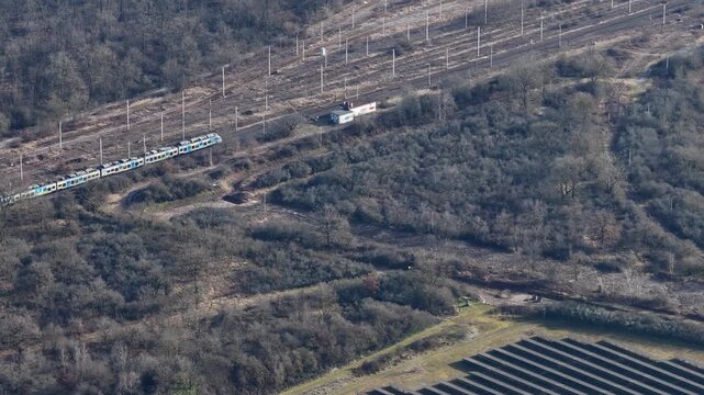 Aerial view of freight and passenger trains moving along parallel tracks, surrounded by bare trees and a small lake, Wittelsheim, Grand Est, France.