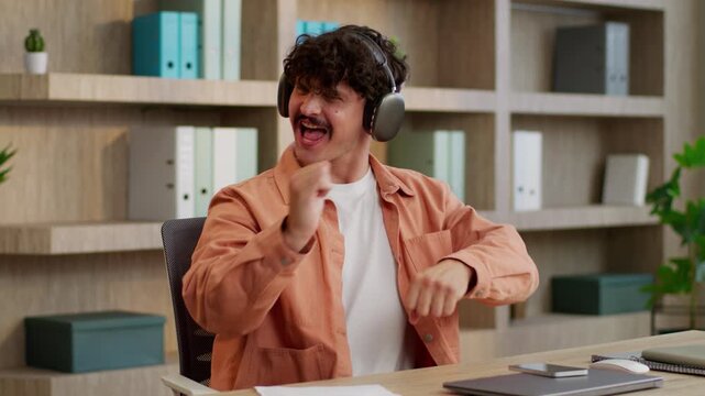 Relaxed Caucasian businessman using mobile phone listening music in headphones dancing business man male guy manager with smartphone wearing earphones dance listen audio funny move at office table