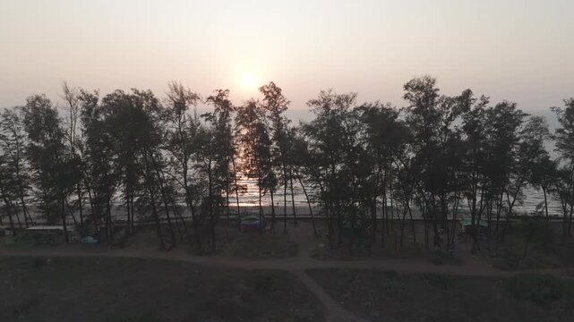 Aerial drone view of Querim Beach at sunset from the Tiracol River, showing a silhouetted coastline with a row of trees, then panning to a glowing sunset over the Arabian Sea