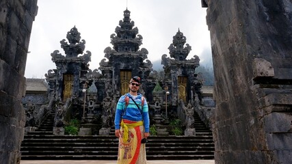 Male tourist in a colorful sarong traditional head covering posing before an ornate stone temple in Bali. Wide shot captures the grand, intricate architecture gray sky. Travel cultural exploration.