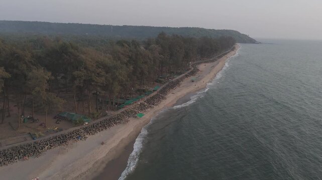 Aerial drone view of Querim Beach, North Goa, revealing a long sandy coastline with scattered rocky outcrops, rows of trees, and the Arabian Sea under soft winter evening light