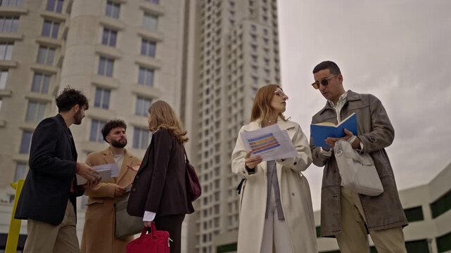 A group of diverse professionals conducts an impromptu outdoor business meeting, intently discussing strategies and reviewing documents against a backdrop of modern city buildings.