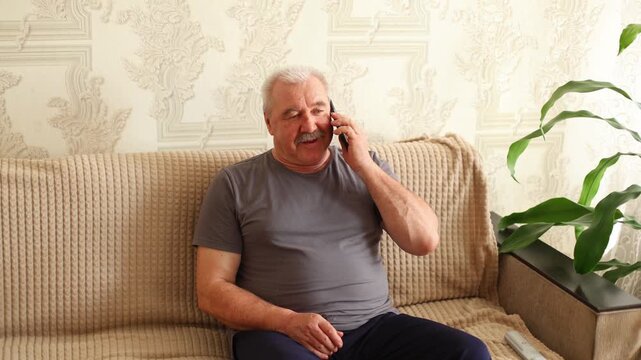 An older man with a mustache sits on a beige sofa, talking on a mobile phone, calm expression, patterned wallpaper, plant nearby.