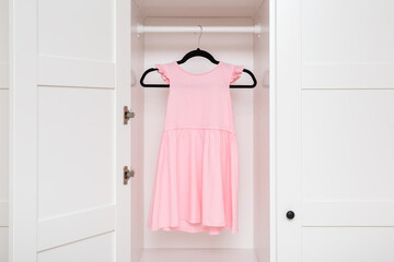 Pastel pink summer child dress hanging on black hanger inside white wooden wardrobe at home room. Closeup. Little girl clothes. Front view.