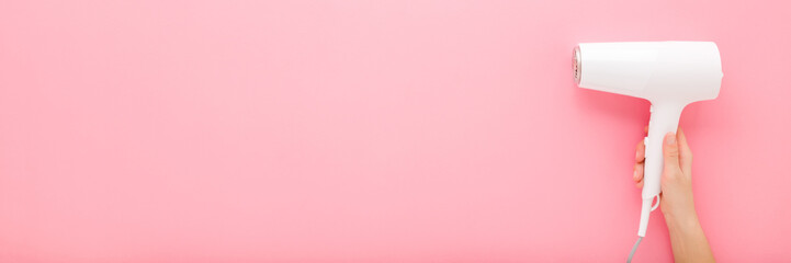 Young adult woman hand holding and showing new white hair dryer on light pink table background. Pastel color. Closeup. Wide banner. Empty place for text. Top down view.