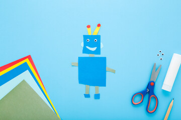 Created robot shape. Scissors, glue stick and colorful application paper on light blue table background. Pastel color. Little child handcraft. Closeup. Top down view.