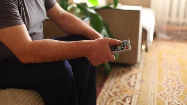 An older man with gray hair and a mustache sits on a sofa, holding a remote, gazing off screen, soft light, patterned wall, green plant in foreground.