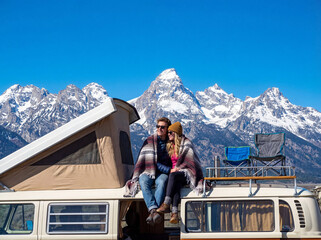 A young couple in love sit on a camper, admiring the view of the mountains and freedom