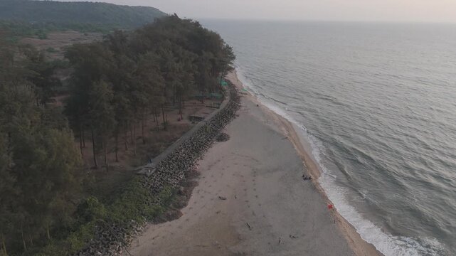 Aerial drone view of Querim Beach, North Goa, revealing a long sandy coastline with scattered rocky outcrops, rows of trees, and the Arabian Sea under soft winter evening light