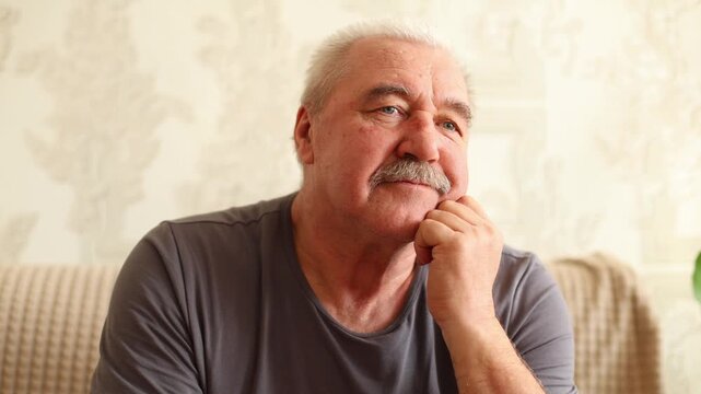 An older man sits thoughtfully on a sofa, resting his chin on his hand, soft lighting, patterned wall, and green leaves frame the quiet, reflective moment.