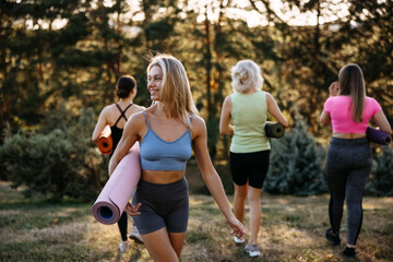 Smiling young woman carrying a yoga mat while walking outdoors with a group of women in a park. Active lifestyle, wellness, fitness, friendship and healthy living concept.