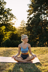 Calm athletic woman sitting in meditation pose outdoors at sunset, practicing mindfulness and relaxation on a yoga mat in a peaceful forest park.