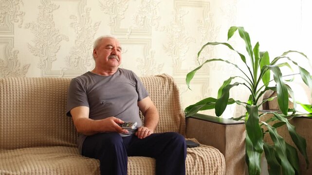 An older man with a mustache sits on a beige sofa, holding a remote, gazing thoughtfully, soft light, patterned wallpaper, and a houseplant frame the scene.