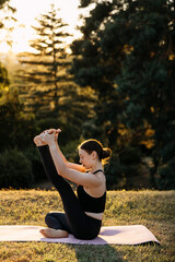 Woman practicing seated balance yoga pose while holding raised leg outdoors at sunset. Flexibility, core strength and mindful movement in nature.