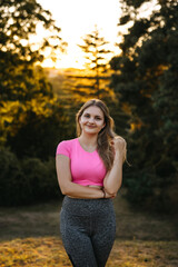 Smiling young woman in pink sports top standing outdoors at golden hour, arms crossed, confident and relaxed fitness lifestyle portrait in nature.
