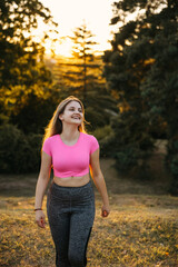 Smiling young woman in pink sports top walking outdoors at sunset. Active lifestyle, fitness and wellness concept with natural light and park background.