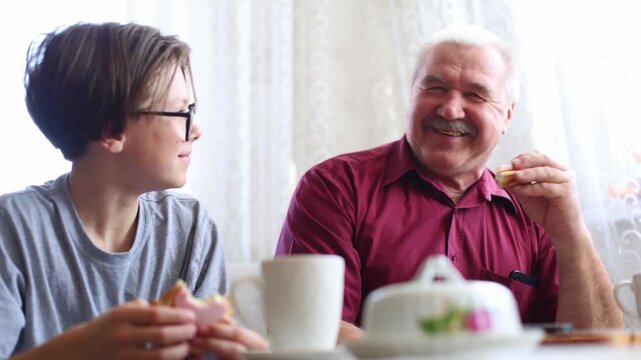 An older man and a boy share breakfast, laughing and playfully high fiving over sandwiches, warm, joyful intergenerational moment at a sunlit table.