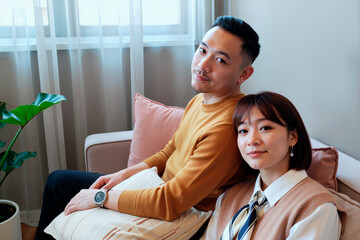 Portrait of young Asian man and young Asian woman sitting together on sofa looking at camera, both relaxed and smiling slightly, casual indoor setting with natural light