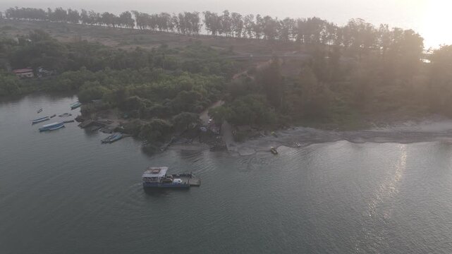 Aerial drone view of Querim Beach at sunset from the Tiracol River, showing a silhouetted coastline with a row of trees, calm water, and a stationary ferry standing in the river