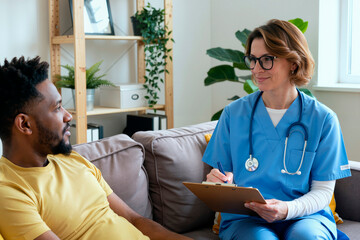 Adult female doctor wearing scrubs sitting on sofa holding clipboard interviewing Black young man during medical consultation in home setting