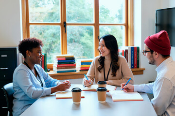 Multiethnic group of young women and young man sitting at table discussing ideas and writing in notebooks with coffee cups and books in background