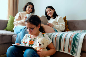 Woman and teenager sitting on sofa using digital tablets while smiling, young girl with glasses holding teddy bear and tablet sitting in foreground focused on screen