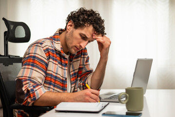 Serious young businessman thinking, taking notes and using laptop while working remotely at his cozy home.