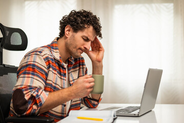 Overworked young businessman having a headache and drinking coffee while working on laptop at his home.	