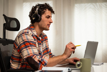 Modern young man using wireless headphones and laptop while participating in e-learning session from the comfort of his home.	