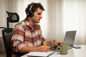 Focused young man wearing wireless headphones, working on his laptop at home while taking notes on a notebook.