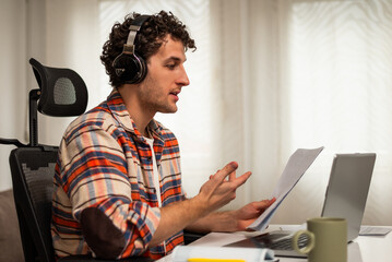 Confident young man with wireless headphones talking and gesturing during a video call on laptop while sitting in his cozy home.	