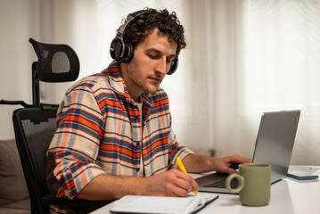 Focused young man wearing wireless headphones, working on his laptop at home while taking notes on a notebook.	