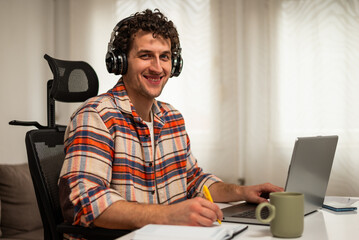 Portrait of happy young man wearing wireless headphones, working on his laptop at home while taking notes on a notebook.