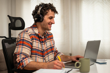 Happy young man wearing wireless headphones, working on his laptop at home while taking notes on a notebook.