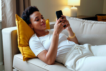 Young Black woman reclining on sofa using smartphone, smiling while interacting with device, relaxed posture, head resting on yellow cushion, casual home environment