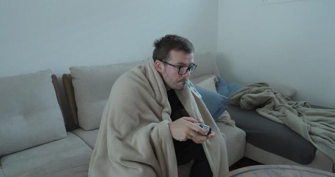 Relatable stock footage showing a sick man wrapped in a blanket, holding a game controller while resting on a sofa at home, symbolizing illness, recovery, boredom, and coping with sickness through gam