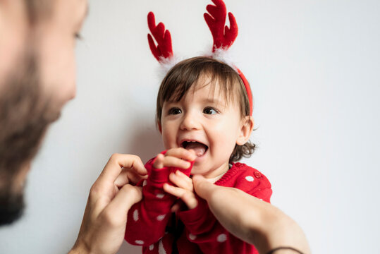 Portrait of laughing baby girl with reindeer antlers headband getting dressed by her father