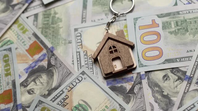 Property Investment and Home Ownership Concept. A wooden house shaped keychain with silver keys lying on a background of scattered dollar bills